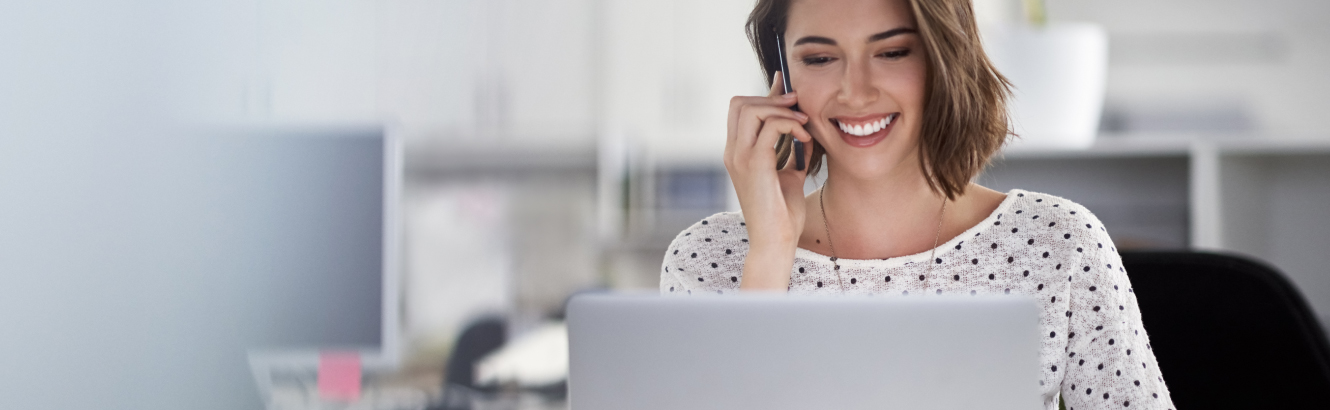 A happy, young woman at work on the phone while browsing her laptop